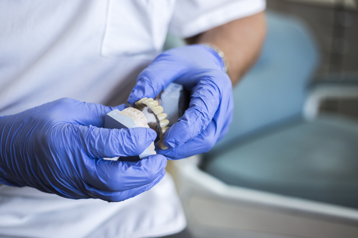 Zahnärztliche Hände mit blauen Handschuhen halten ein Modell mit eingesetztem Zahnersatz im Behandlungsraum.