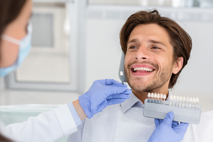 Dentist checking on male patient teeth color Patient im Behandlungsstuhl zeigt fröhlich eine Farbskala für Zahnaufhellung und lacht dabei herzlich.