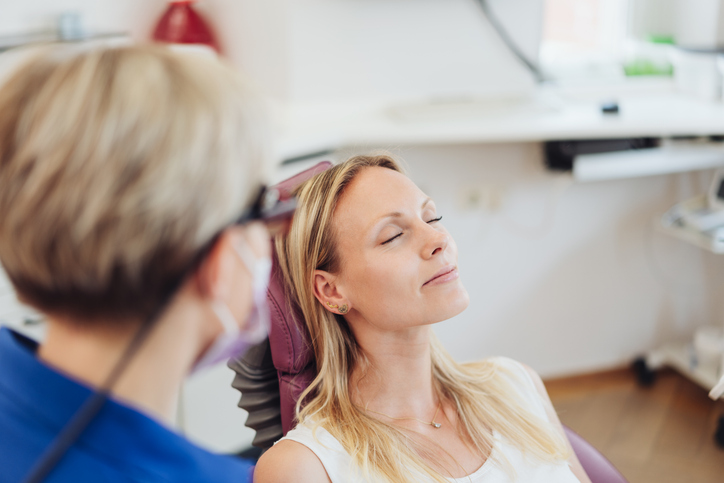 Woman relaxing with her eyes close at the dentist Patientin lehnt sich entspannt im Zahnarztstuhl zurück. Behandelnde Zahnärztin bereitet sich auf Untersuchung vor.