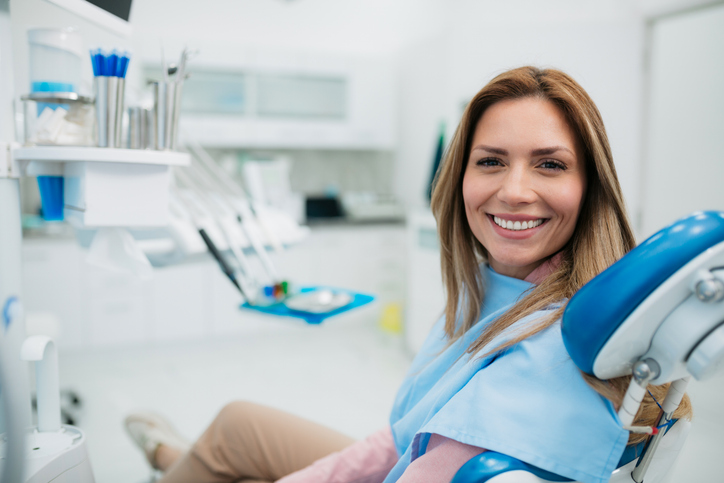 Happy woman visiting a dentist office Patientin sitzt auf einem Behandlungsstuhl, lächelt freundlich in Kamera. Im Hintergrund sieht man den Behandlungsraum.