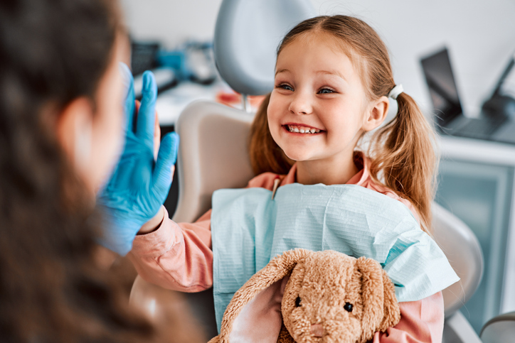 Children's dentistry. Mädchen sitzt lachend im Zahnarztstuhl, hält ein Kuscheltier im Arm und schaut zur behandelnden Zahnärztin.