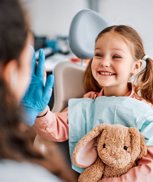 Children's dentistry. Mädchen sitzt lachend im Behandlungsstuhl, hält ein Kuscheltier und schaut zur freundlichen Zahnärztin.