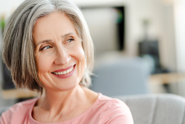 Smiling confident senior woman sitting on comfortable sofa at home. Happy female looking away Ältere Patientin mit grauen Haaren sitzt entspannt auf einem Sofa und lächelt zufrieden in die Kamera.